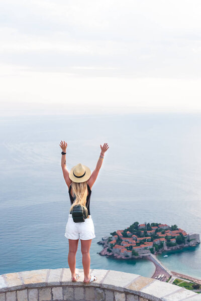 Вид сзади женщины, стоящей с поднятыми руками и смотрящей на остров Стейнт Стефен в Адриатическом море, Будва, Монтенегро
