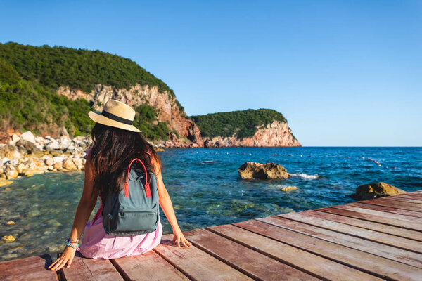 Вид сзади женщины с рюкзаком, сидящей на мосту у моря в Будве, Монтенегро
