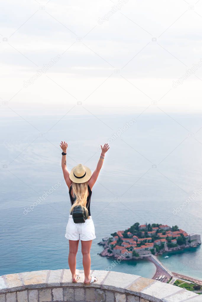 back view of woman standing with raised hands and looking at saint stephen island in Adriatic sea, Budva, Montenegro