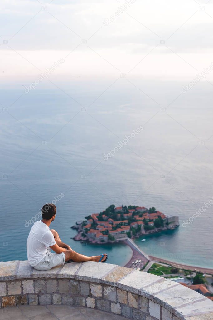 man sitting on viewpoint and looking at island of Sveti Stefan with hotel resort in Adriatic sea, Budva, Montenegro