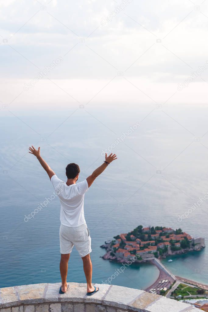 back view of man with raised hands looking at island of Sveti Stefan with hotel resort in Adriatic sea, Budva, Montenegro
