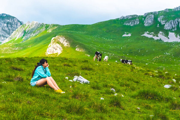 beautiful woman sitting near cows grazing on green valley in Durmitor massif, Montenegro