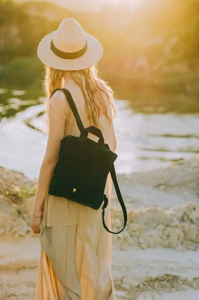日光湖近くに立っているバックパックと帽子でブロンドの女の子の背面図 — ストック写真