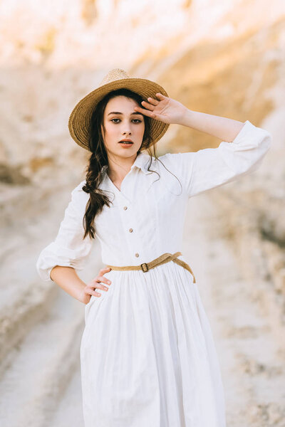 beautiful stylish girl in white dress and straw hat posing in sandy canyon