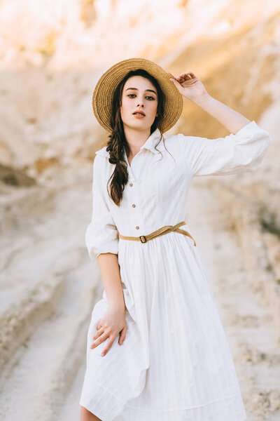 очаровательная стильная девушка позирует в белом платье и соломенной шляпе в песчаном каньоне
