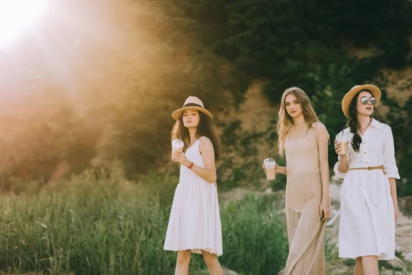 charming girls in straw hats holding coffee latte and walking in nature with back light