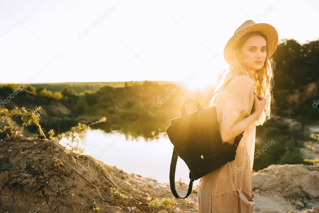 stylish elegant girl in straw hat posing with backpack near lake with back light