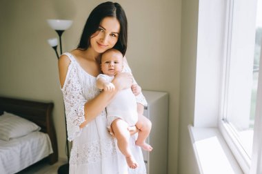 gülümseyen genç anne çocuğunu evde pencere tutarak ve kameraya bakıyor