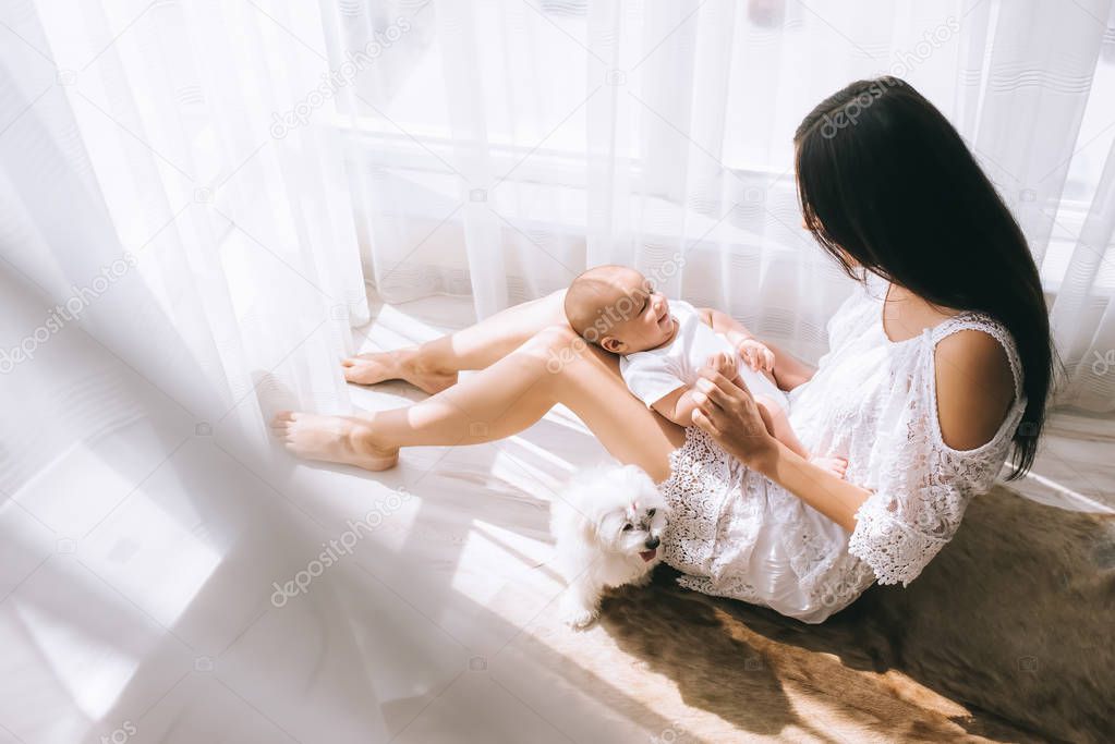happy young mother playing with her baby while sitting on floor at home with bichon dog