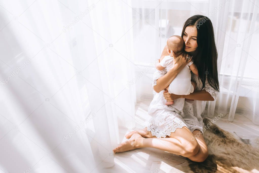 happy young mother embracing her baby boy while sitting on floor at home