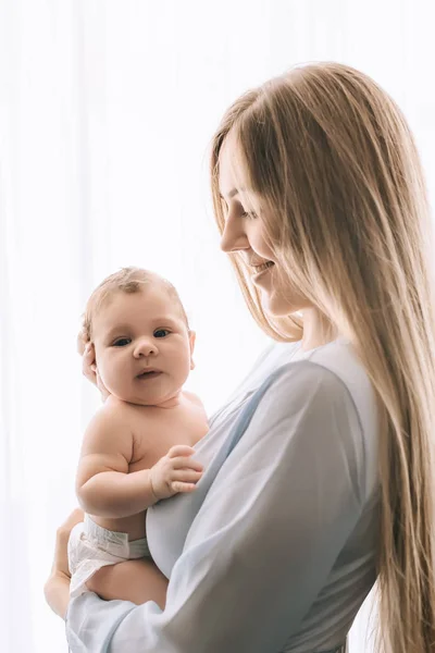 Zijaanzicht Van Lachende Moeder Uitvoering Baby Jongetje Voor Gordijnen Thuis — Stockfoto