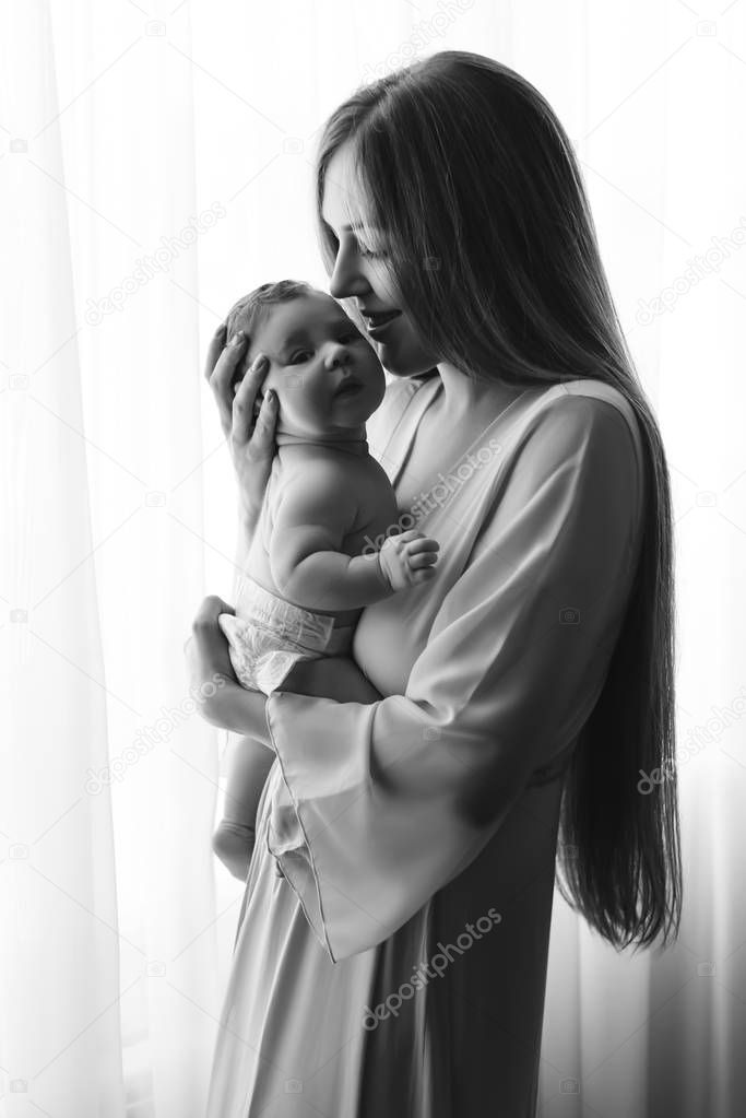 black and white picture of mother carrying little baby boy in front of curtains at home