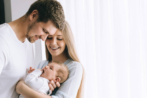 cheerful family carrying little baby boy in front of curtains at home