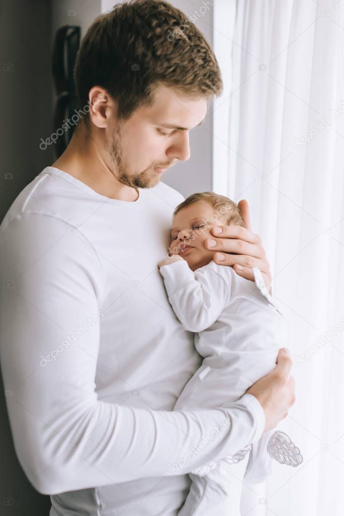 handsome father carrying little baby boy in front of curtains at home