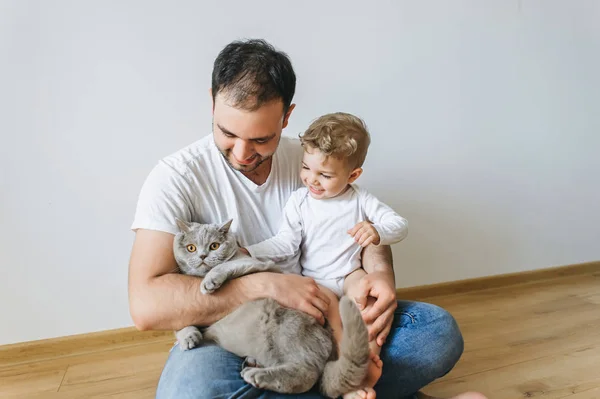 Padre sorridente che tiene in braccio il piccolo figlio in body e il gatto pantaloncini grigio britannico in mano a casa — Foto stock