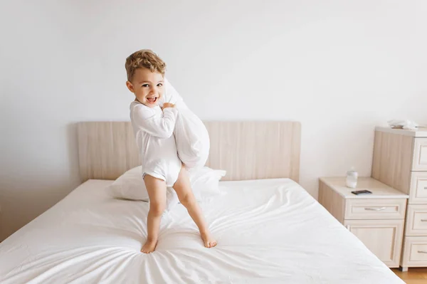 Adorável criança menino em terno branco brincando com travesseiro na cama em casa — Fotografia de Stock