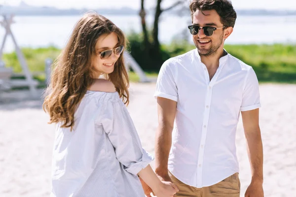 Sorrindo casal em óculos de sol de mãos dadas na praia da cidade arenosa — Fotografia de Stock