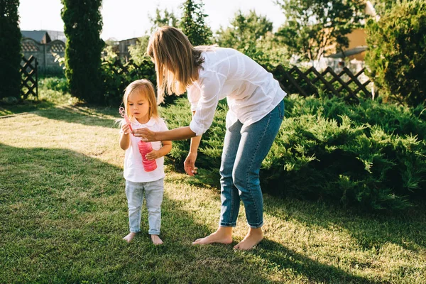 Madre ayudando a la pequeña hija soplando burbujas de jabón en el patio trasero - foto de stock