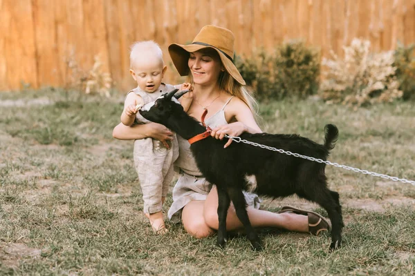 Mãe sorridente em chapéu e pequeno filho acariciando cabra doméstica na grama verde no campo — Fotografia de Stock