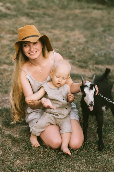 Mãe sorridente no chapéu, pequeno filho e cabra doméstica na grama verde no campo — Fotografia de Stock