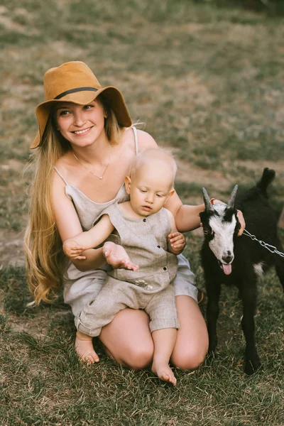 Smiling mother in hat, little son and domestic goat on green grass at countryside — Stock Photo