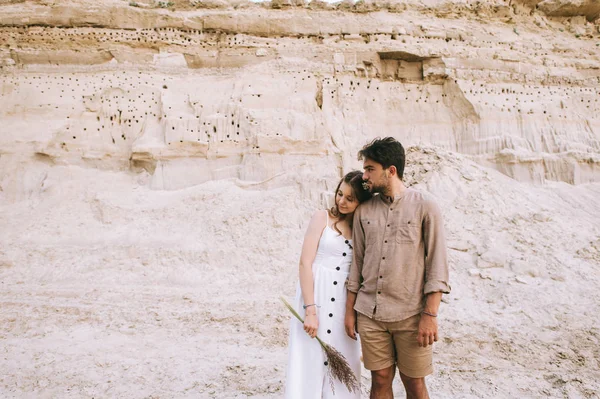 Jovem menina atraente em vestido branco segurando buquê floral e abraçando namorado no desfiladeiro de areia — Fotografia de Stock