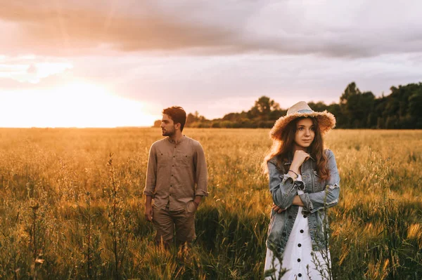 Elegante novia en sombrero de paja posando con el novio en el prado de verano al atardecer con retroiluminación - foto de stock