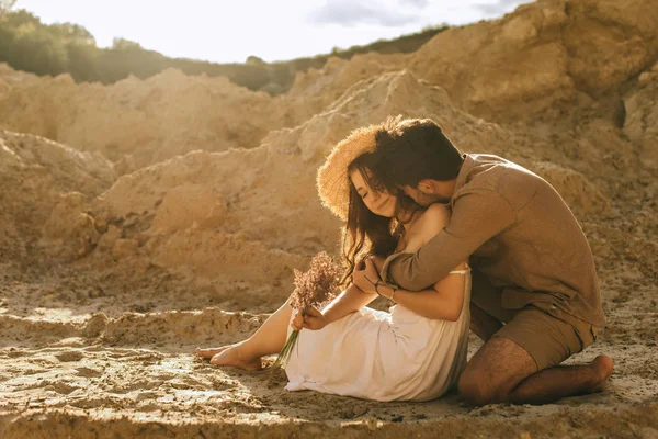 Happy man hugging girlfriend in straw hat with floral bouquet in sand canyon — Stock Photo