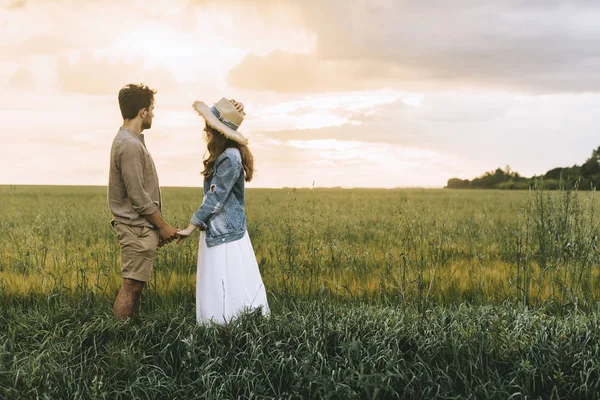 Stylish couple holding hands on green field and looking at sunset — Stock Photo