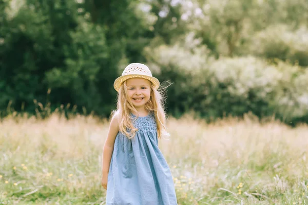 Adorable enfant souriant blonde en robe et chapeau de paille dans le champ — Photo de stock