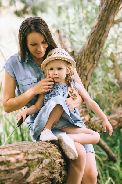 Madre feliz y adorable hija rubia sentada en el tronco del árbol - foto de stock