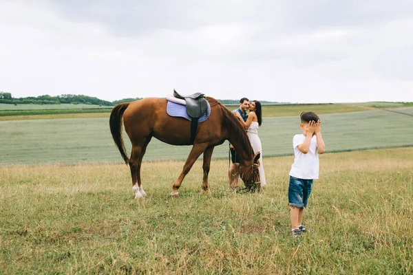 Padres abrazándose cerca de caballo, hijo cubriendo los ojos en el campo - foto de stock