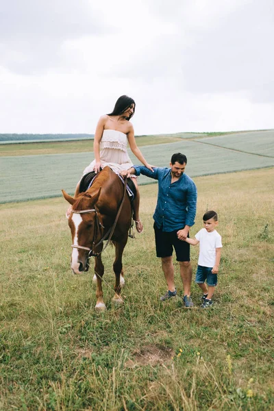 Mère chevauchant cheval brun, père et fils debout sur le terrain — Photo de stock