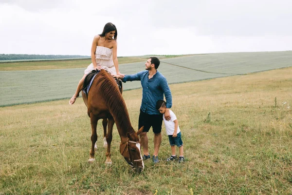 Mãe equitação marrom cavalo, pai e filho de pé no campo — Fotografia de Stock