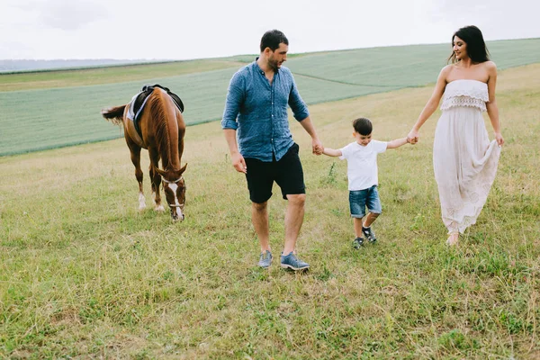 Parents and son holding hands and walking near brown horse on field — Stock Photo