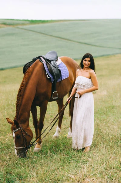 Schöne Frau, die mit braunem Pferd auf dem Feld steht und in die Kamera schaut — Stockfoto