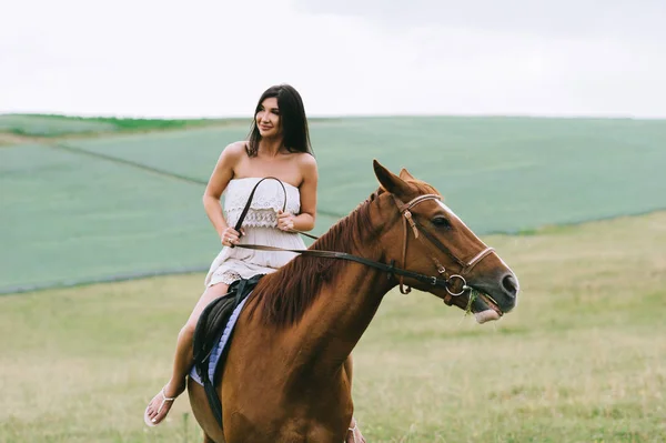 Bela mulher montando cavalo marrom no campo verde — Fotografia de Stock