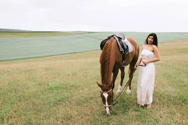 Hermosa mujer de pie cerca de caballo marrón en el campo - foto de stock