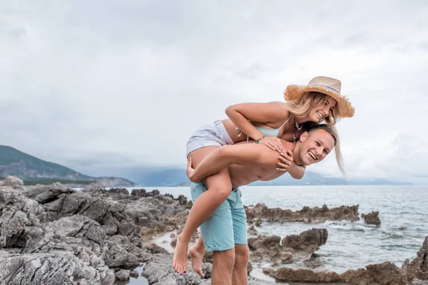 Feliz joven pareja piggybacking y riendo en rocosa playa en montenegro - foto de stock