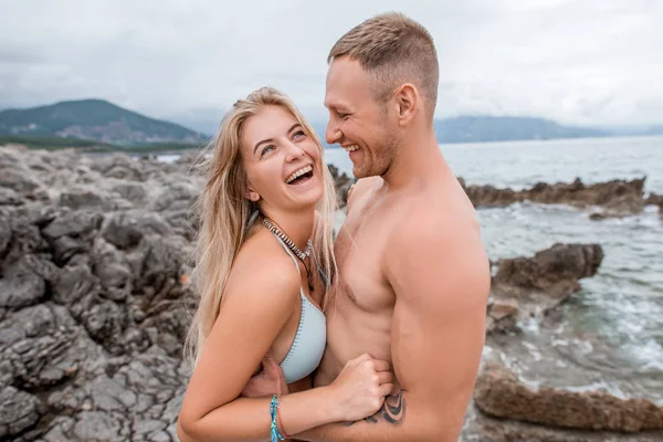 Vista lateral de feliz pareja joven abrazando y riendo en la playa rocosa en montenegro - foto de stock