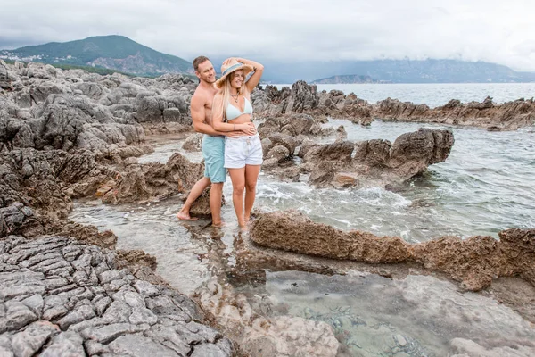 Счастливая молодая пара обнимается и смотрит в сторону, стоя вместе на скалистом пляже в Черногории — стоковое фото