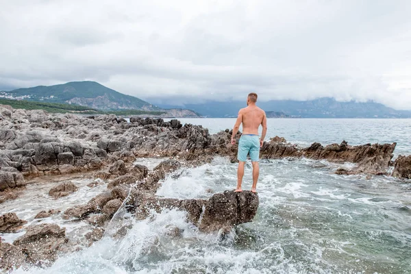 Vista trasera del joven musculoso guapo parado en la playa rocosa y mirando majestuosa vista al mar en montenegro - foto de stock