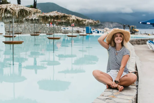 Bela feliz jovem mulher em chapéu de palha sentado perto da piscina e sorrindo para a câmera, monten= — Fotografia de Stock