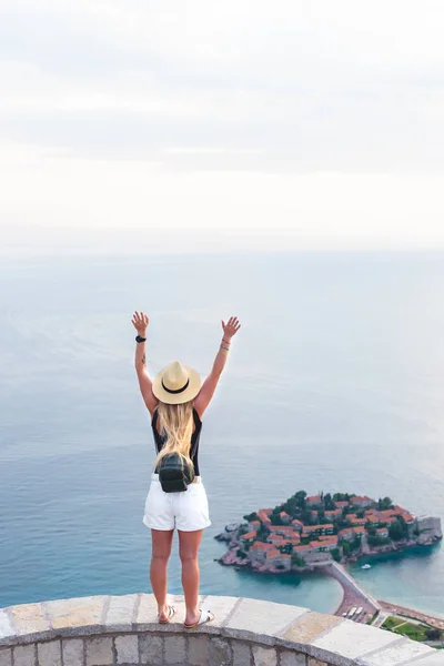 Vista posteriore della donna in piedi con le mani alzate e guardando santo stephen isola nel mare Adriatico, Budva, Montenegro — Foto stock