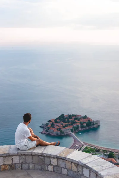 Hombre sentado en el mirador y mirando a la isla de Sveti Stefan con hotel resort en el mar Adriático, Budva, Montenegro - foto de stock