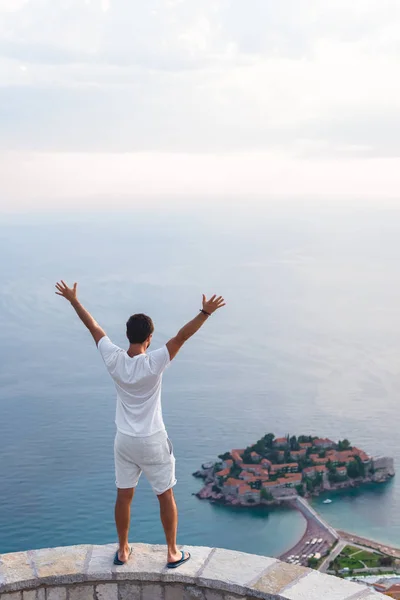 Visão traseira do homem com as mãos levantadas olhando para a ilha de Sveti Stefan com hotel resort no mar Adriático, Budva, Montenegro — Fotografia de Stock