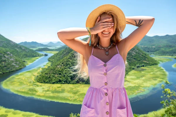 Femme souriante en robe rose et chapeau couvrant les yeux près de la rivière Crnojevica (Rijeka Crnojevica) au Monténégro — Photo de stock
