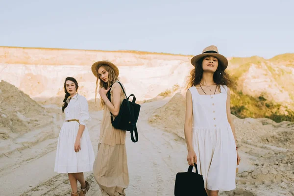 Chicas bonitas en vestidos elegantes y sombreros de paja caminando con mochilas en cañón de arena - foto de stock