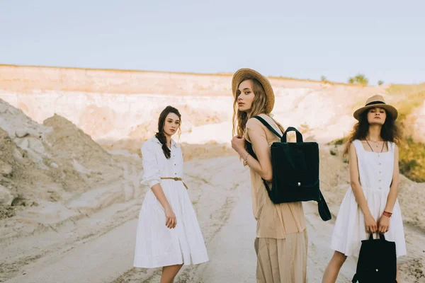 Elegantes namoradas elegantes em chapéus de palha andando com mochilas em canyon arenoso — Fotografia de Stock