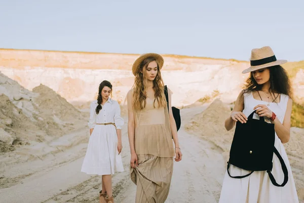 Beautiful stylish girlfriends in straw hats walking with backpacks in sandy canyon — Stock Photo